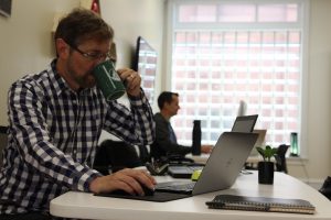 an image of a man close up coworking in williamsport, working at a desk at the Covation Center, drinking a coffee and looking at a computer, with another man in the background doing the same. 